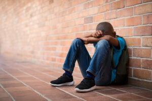 A young boy sitting with his back against a red brick wall, covering his face with his arms